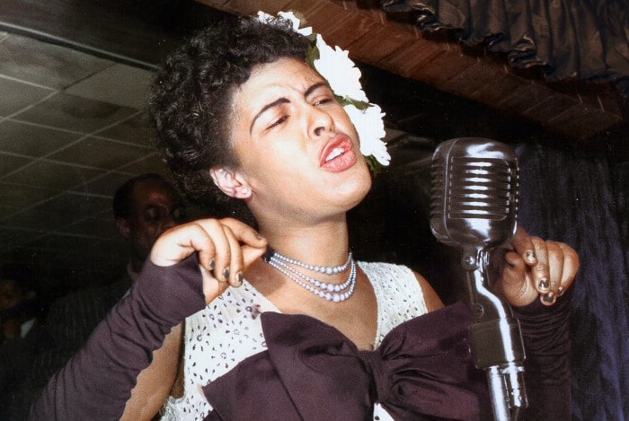 Billie Holiday in ihrem Element auf der Bühne. Sie trägt ein weisses Träger Kleid mit einer lila Schleife vor ihrer Brust.