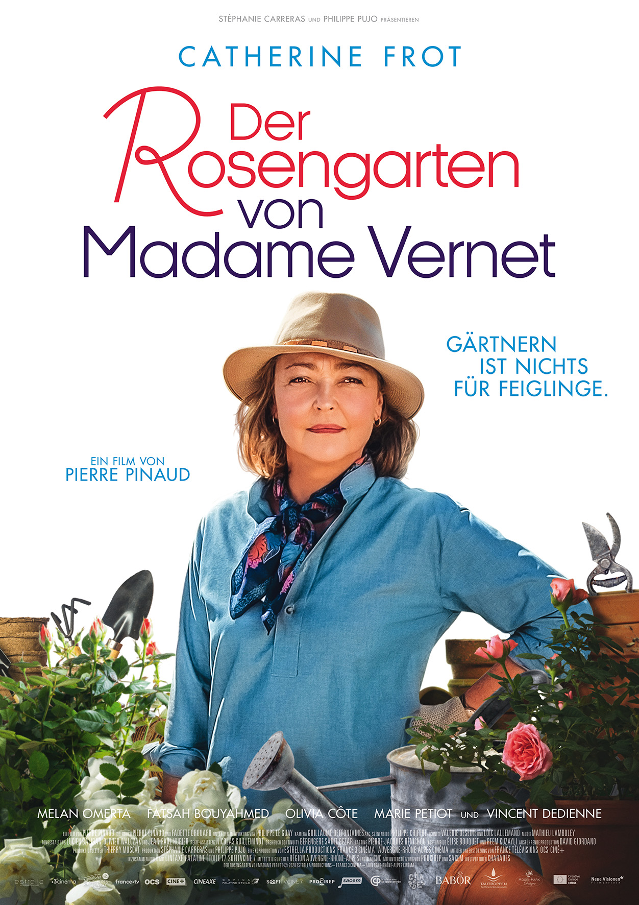 Poster mit Schriftzug Der Rosengarten von Madame Vernet. Schauspielerin Catherine Frot inmmitten von Blumen mit einem Hut auf dem Kopf. Sie trägt ein blaues Hemd und einen leichten Schal um den Hals