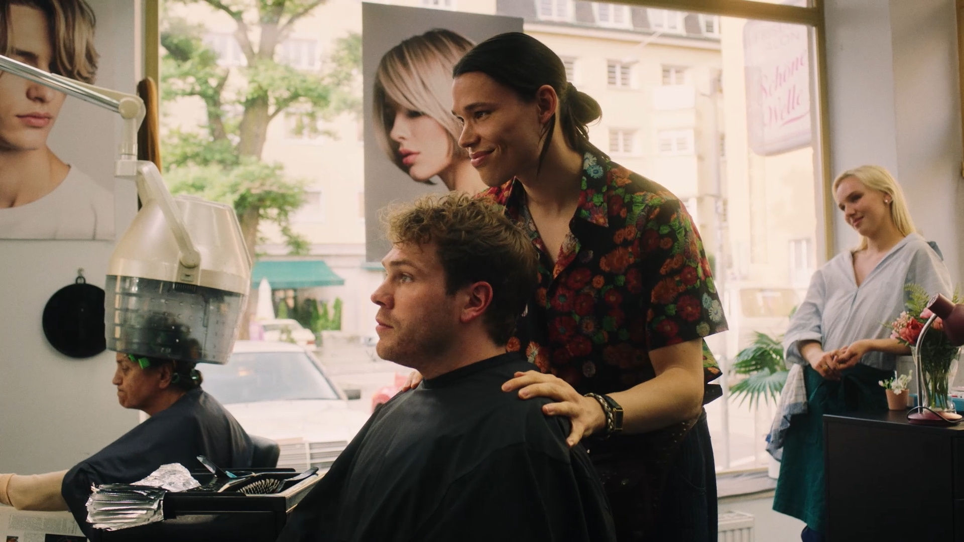 "München Beats - Folge 2": Im Frisörsalon: Andreas Bierwirth (Ben Münchow) sitzt vor John (Helge Mark) auf einem Friseurstuhl. Beide blicken zur Seite. Im Hintergrund steht Linda Bierwirth (Jule Hermann) vor einem großen Glasfenster und beobachtet die beiden. Eine weitere Person (Komparsin) sitzt im Hintergrund unter einer Trockenhaube.
ZDF/ Holly Fink
